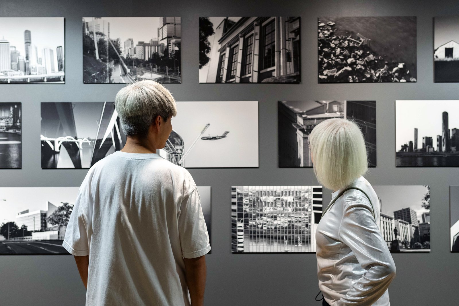 A wall of black and white photos. Two people with their backs to the camera, are wearing white. Their hair too is in shades of white/grey.